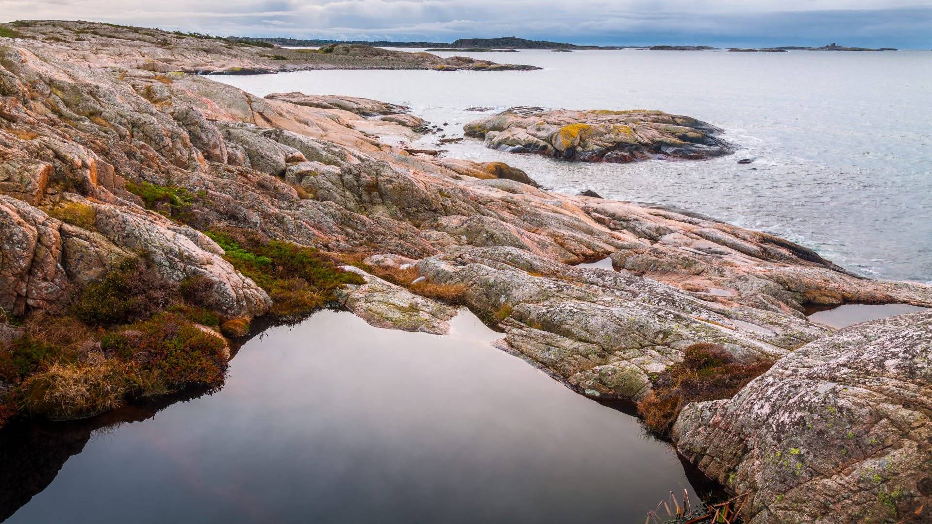 Klippig kustlinje vid Kosterhavets nationalpark med en liten vattensamling i förgrunden och med ett stilla hav och avlägsna öar i bakgrunden.